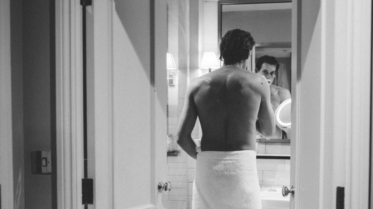 Uomo attraente che applica un rasoio di sicurezza sul viso coperto di schiuma mentre si rade davanti a uno specchio del bagno durante una tradizionale rasatura bagnata